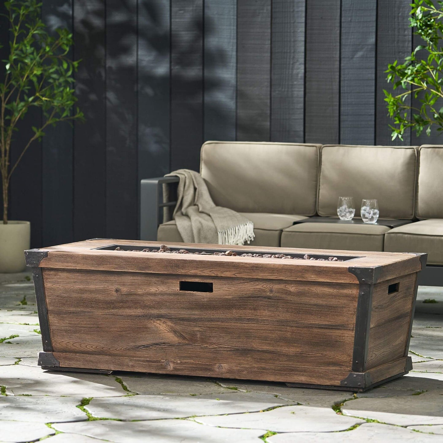 Wooden fire pit table on a patio with a beige sofa and plants in the background.
