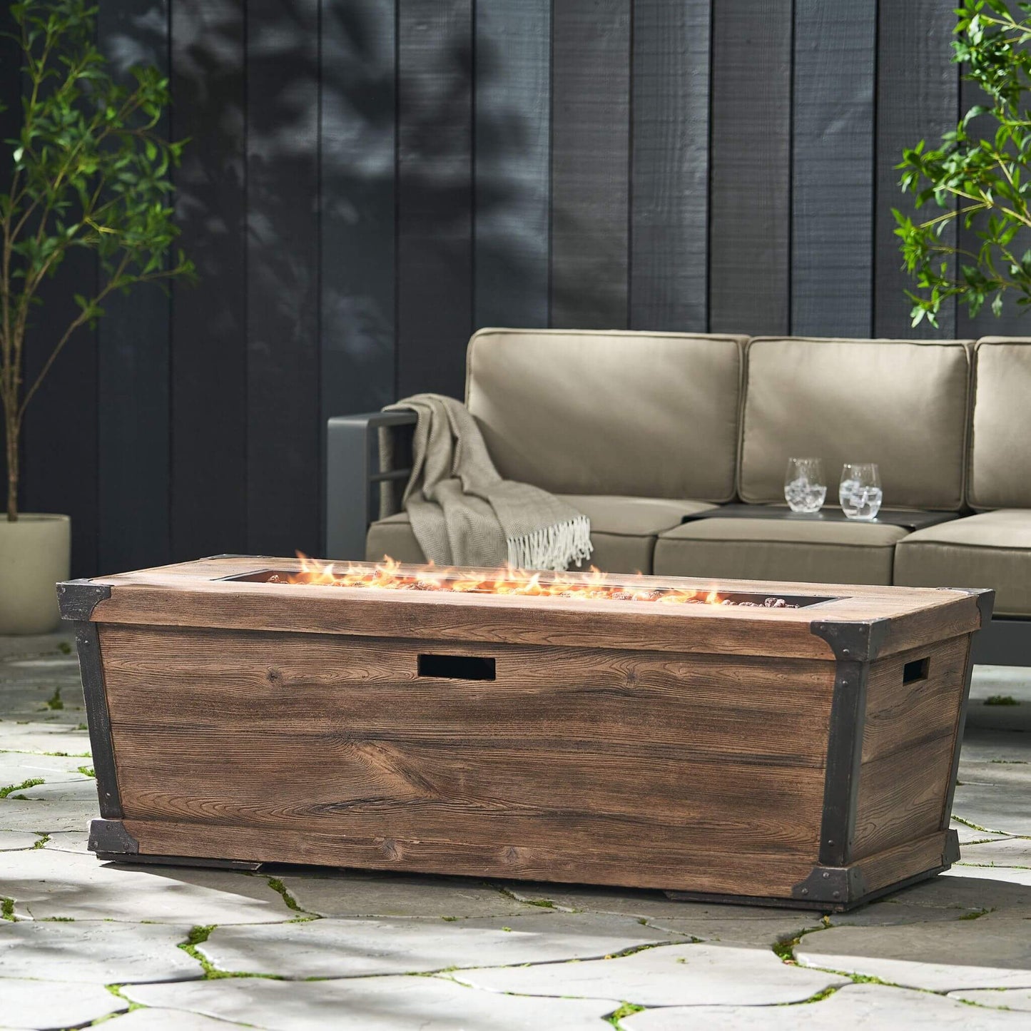Wooden fire pit table with flames on a patio, surrounded by a beige sofa and plants.