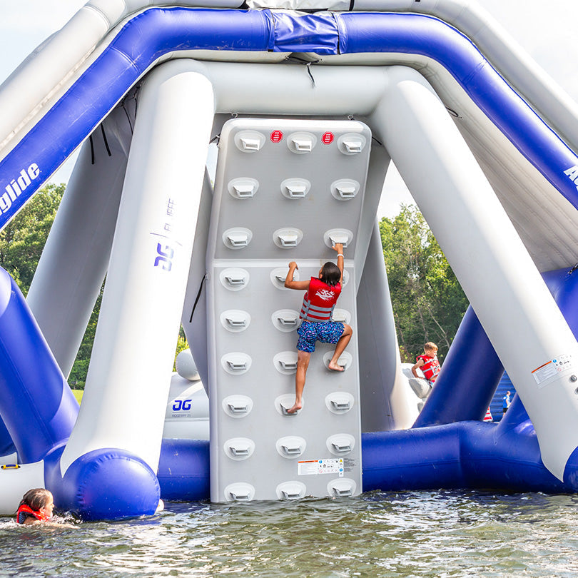 ser climbing the Aquaglide Escalade Summit Climbing Wall, a 16 ft tall aquapark feature, in a lake setting.