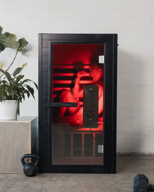 Black sauna with red interior light, plant, and kettlebell on a concrete floor.
