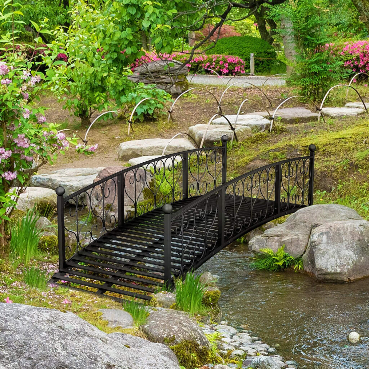 Black metal bridge over a stream in a garden with flowers and rocks