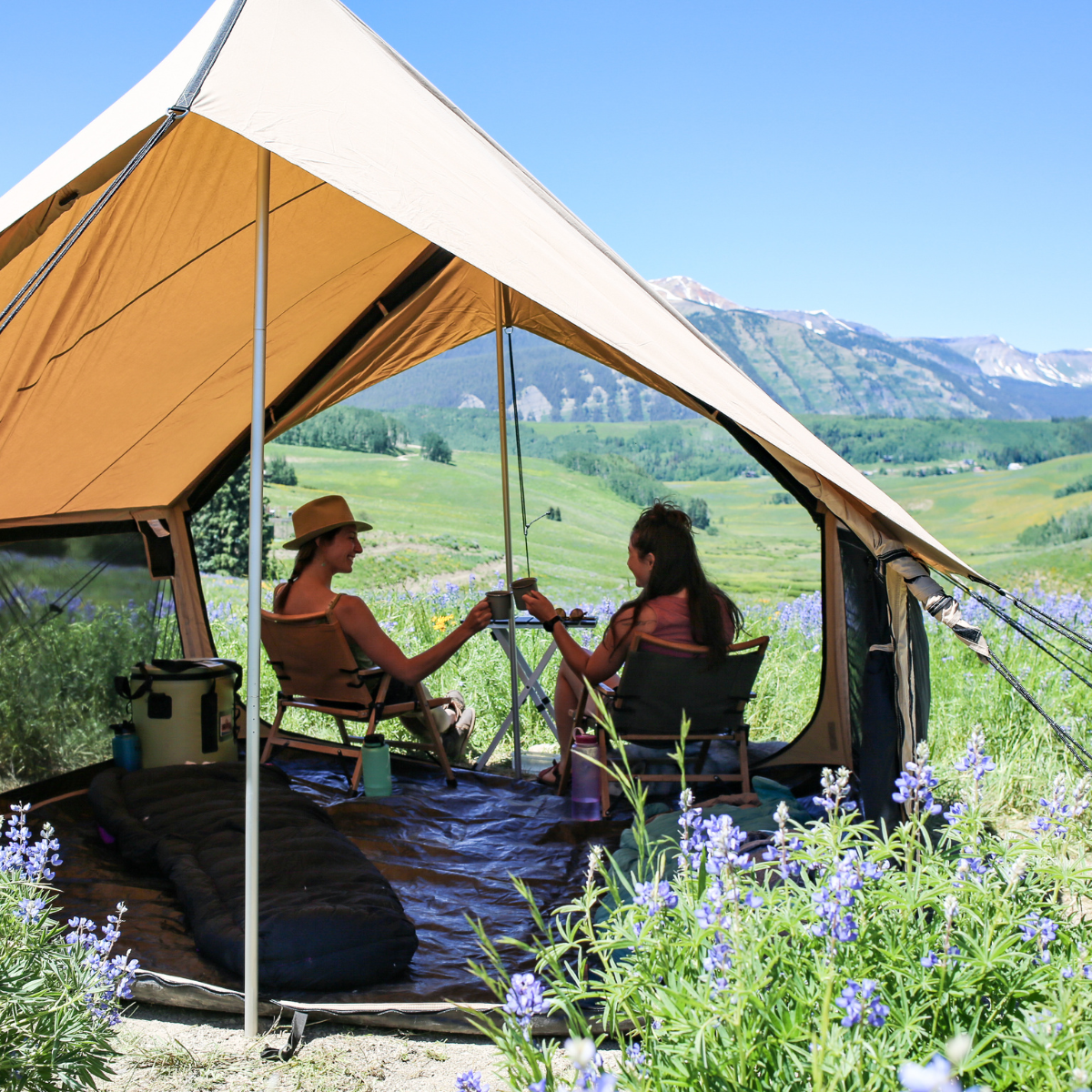 Two people sitting in a tent overlooking a scenic landscape with mountains and fields.