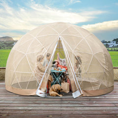 A large beige outdoor igloo garden greenhouse dome tent with a transparent TPU surface. It is set up on a wooden deck with plants inside and a dog lying on the floor.