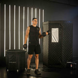 Man holding dumbbells in front of a black sauna box with illuminated vertical lights on the wall.