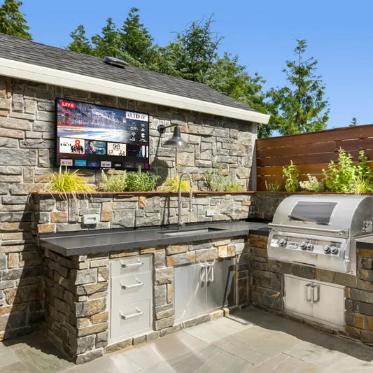 Outdoor kitchen with stone wall, outdoor 4K TV, and grill on a sunny day.