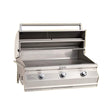 A built-in Fire Magic Choice Multi-User Grill with analog thermometer, featuring three control knobs and a stainless steel construction, displayed against an orange background.
