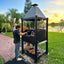 Person grilling outdoors using a large black barbecue grill in a garden setting with trees and a lake in the background.