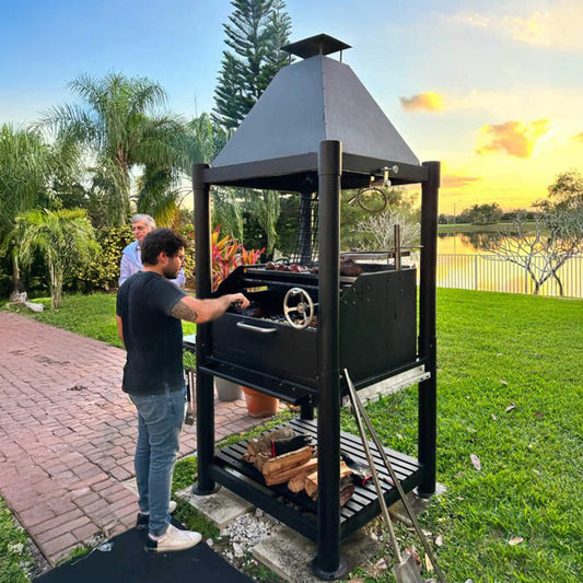 Person grilling outdoors using a large black barbecue grill in a garden setting with trees and a lake in the background.